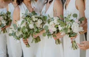 Women in all white sleeveless dresses and jumpsuits, holding bouquets