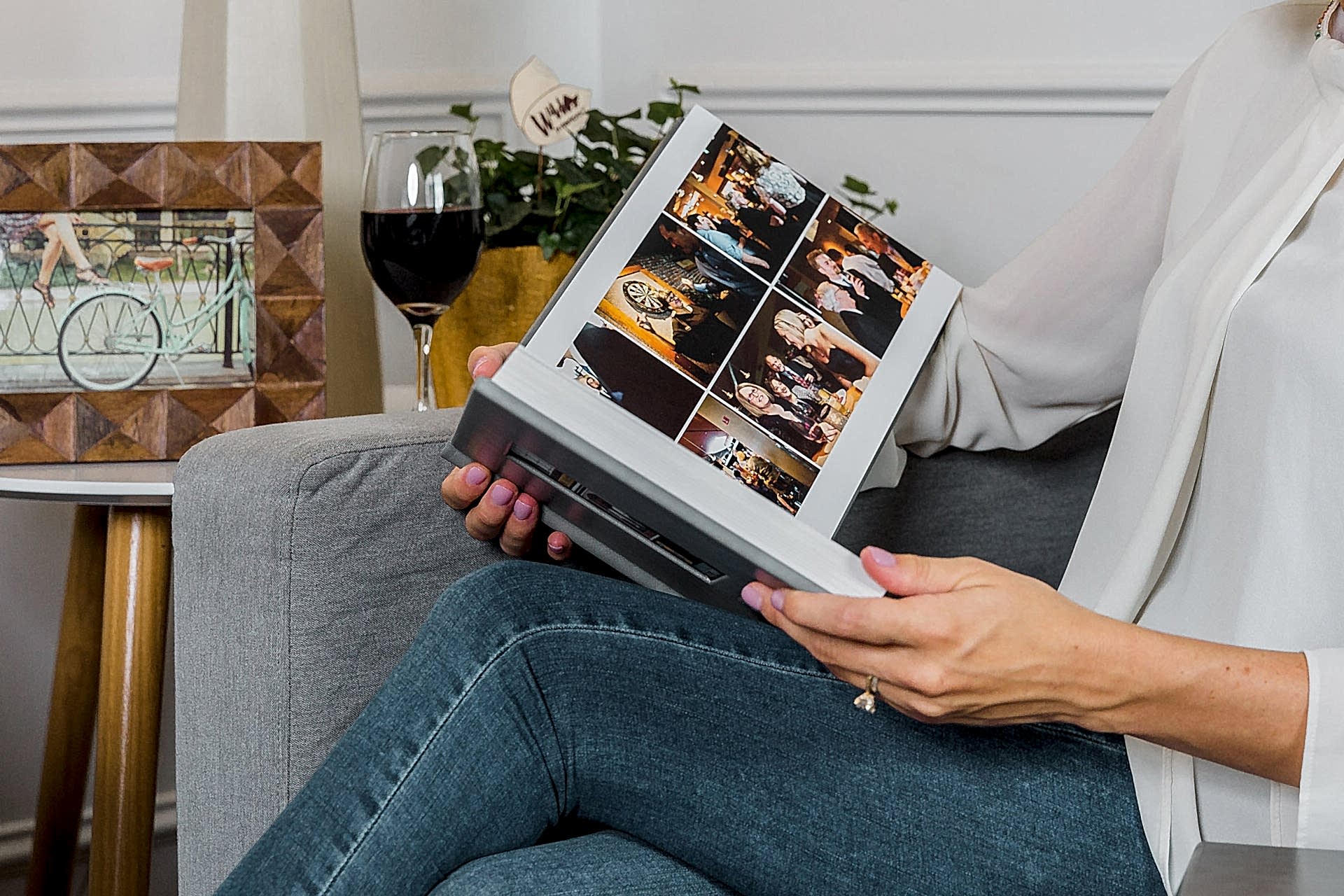 A woman in a white sweater and jeans sits on a couch looking through a grey leather-bound album from Albums Remembered while a glass of red wine sits on the end table beside her