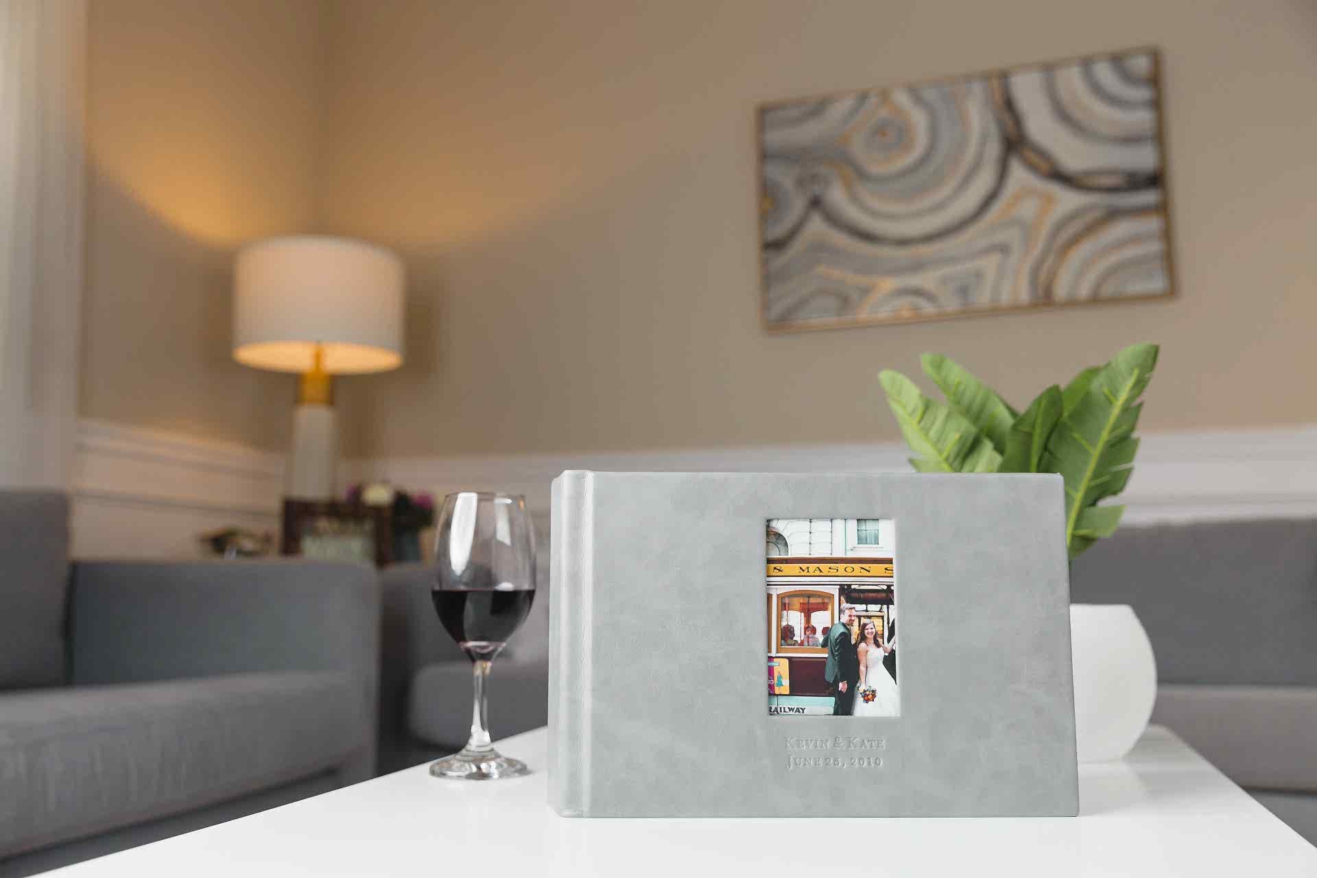 A grey leather-bound wedding album from Albums Remembered sits on the white coffee table of a modern living room alongside a glass of red wine