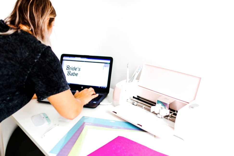 A woman using a laptop to design text to cut next to her Cricut Explore Air 2 Machine from Michael's Weddings