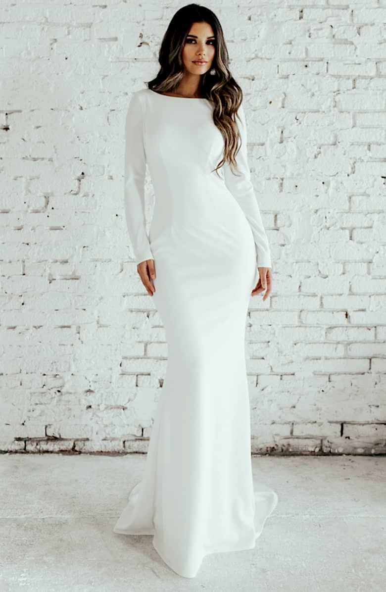 brunette woman wearing a long sleeve wedding gown standing in from of a white brick wall
