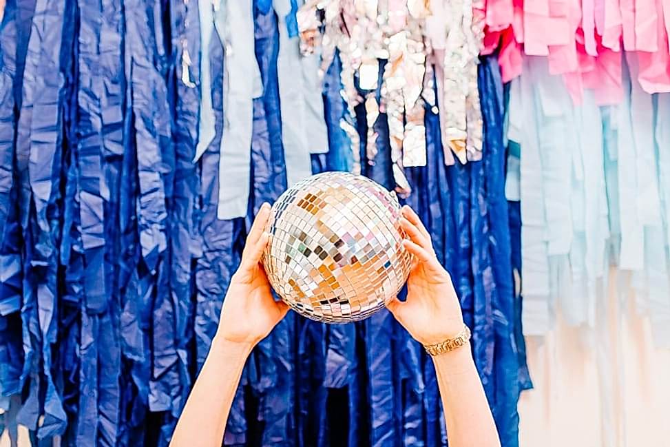 A small disco ball held by a woman's hands in front of a colorful fringe installation at The Compact Camp