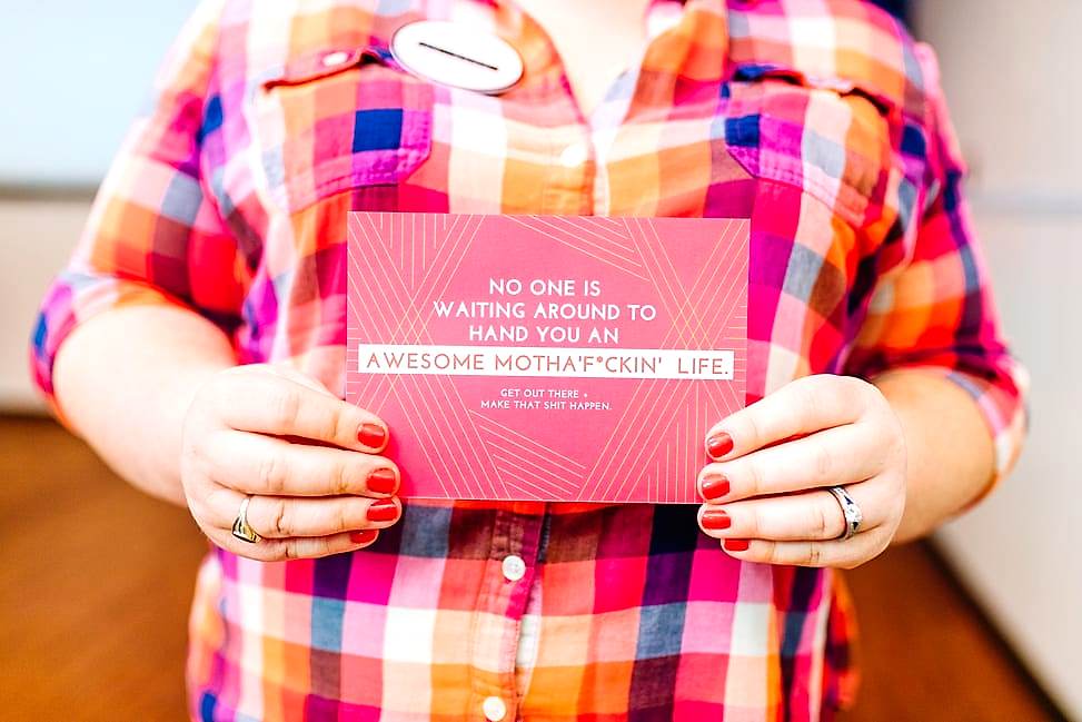 A woman in a brightly colored plaid shirt with red nails holds a pink postcard that reads "No one is waiting around to hand you an awesome mothaf*ckin life. Get out there + make that shit happen.