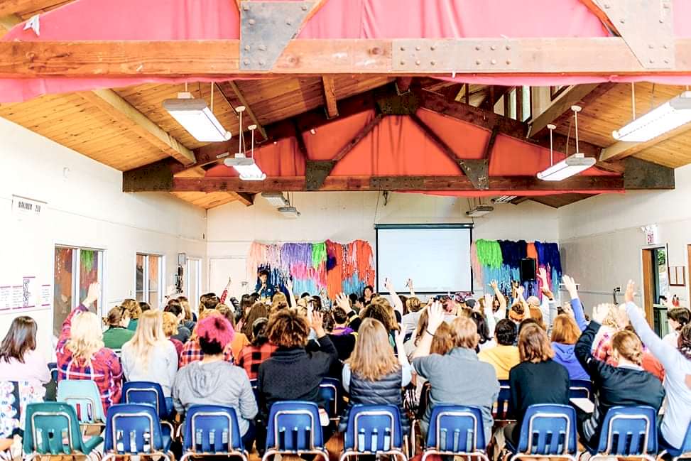 A roomful of women in school-style chairs raise their hands during Cyndie Spiegel's keynote at The Compact Camp