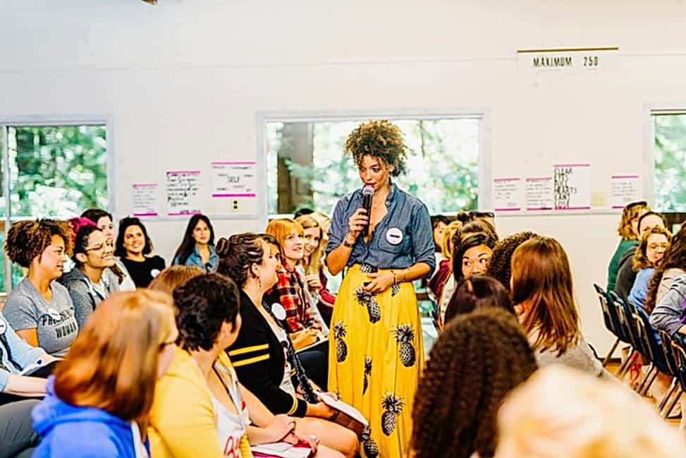 Cyndie Spiegel wearing a yellow pinapple print skirt and chambray button down top holds a microphone and stands in the audience during her keynote speech at The Compact Camp