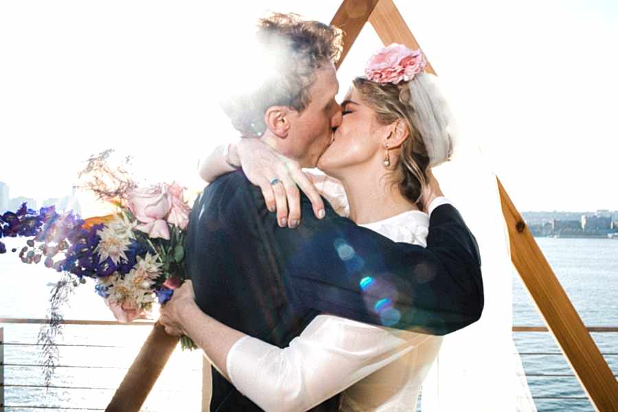 A Corey Torpie photo of two people in wedding kissing in a tight embrace in front of a wooden triangular altar piece, overlooking the water.