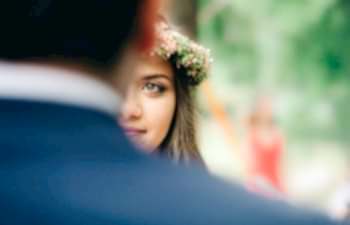A woman with a flower crown, hazel eyes, and brown hair looks forward toward you at the man she is marrying who is out of focus with his back to you, wearing a blue skirt.