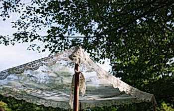 A lace chuppah with a wooden post underneath a green tree in the sunlight