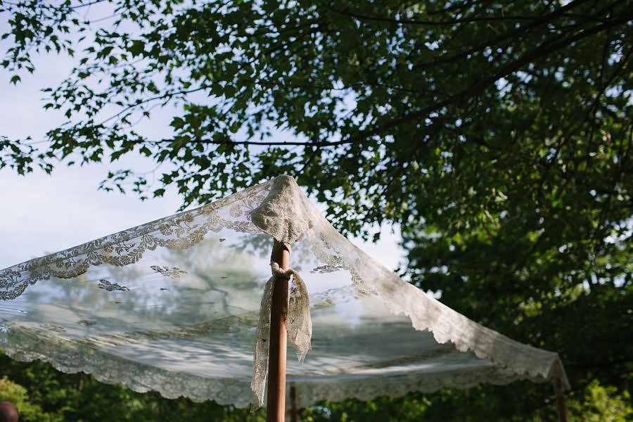 A lace chuppah with a wooden post underneath a green tree in the sunlight