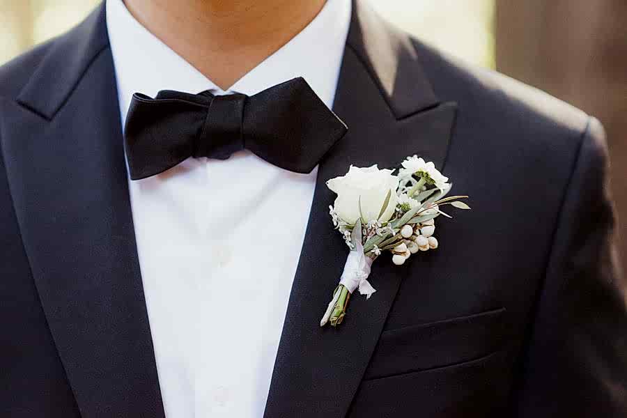 The torso of a man in a black tux with bow tie, wearing a white floral boutonnière.