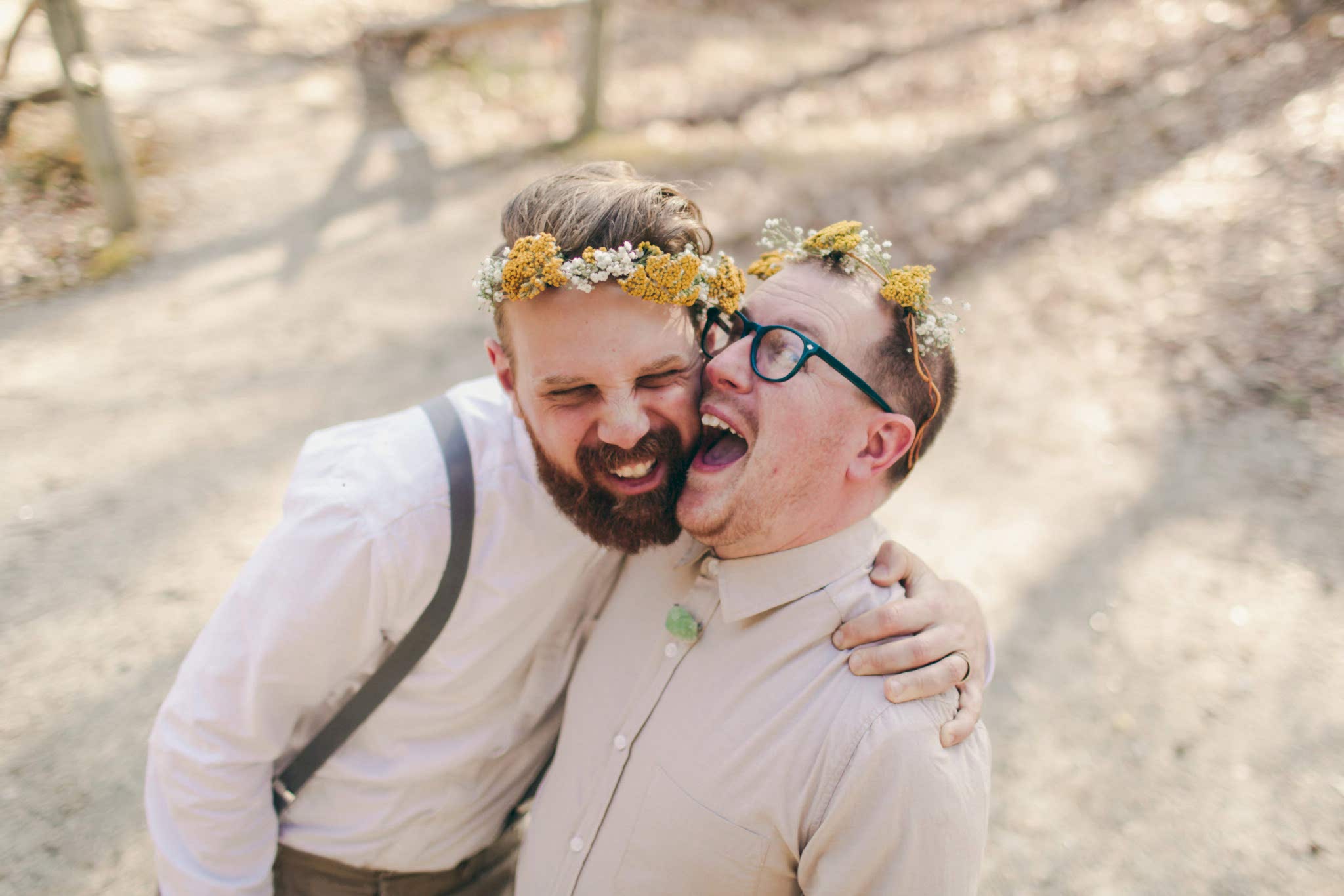 Two grooms in tan shirts and yellow flower crowns laugh and embrace in a photo by Betty Clicker