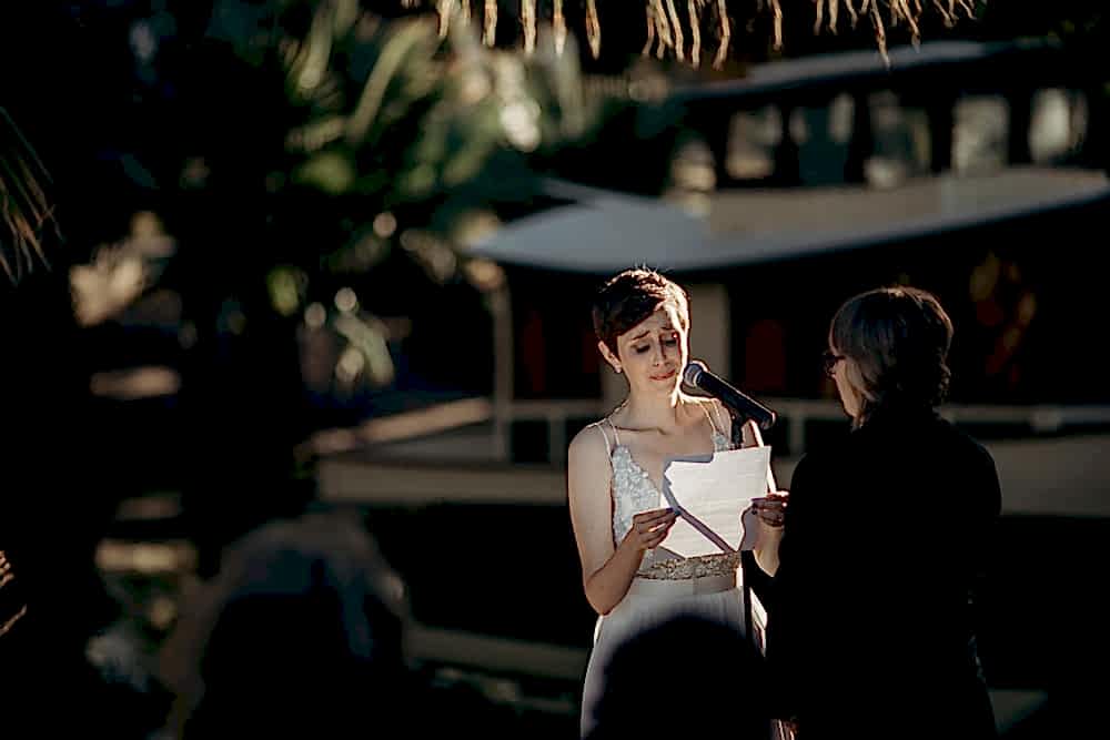 Bride with short hair and beaded gown is tearing up as she faces her partner and reads her vows in a photo by Betty Clicker