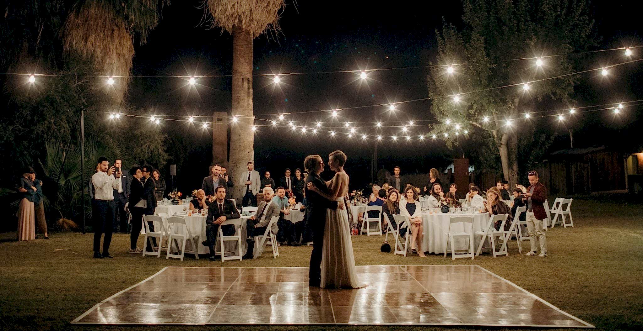 A wedding couple has their first dance on a outdoor dance floor surrounded by palm trees and string lights as their guests look on in a photo by Betty Clicker