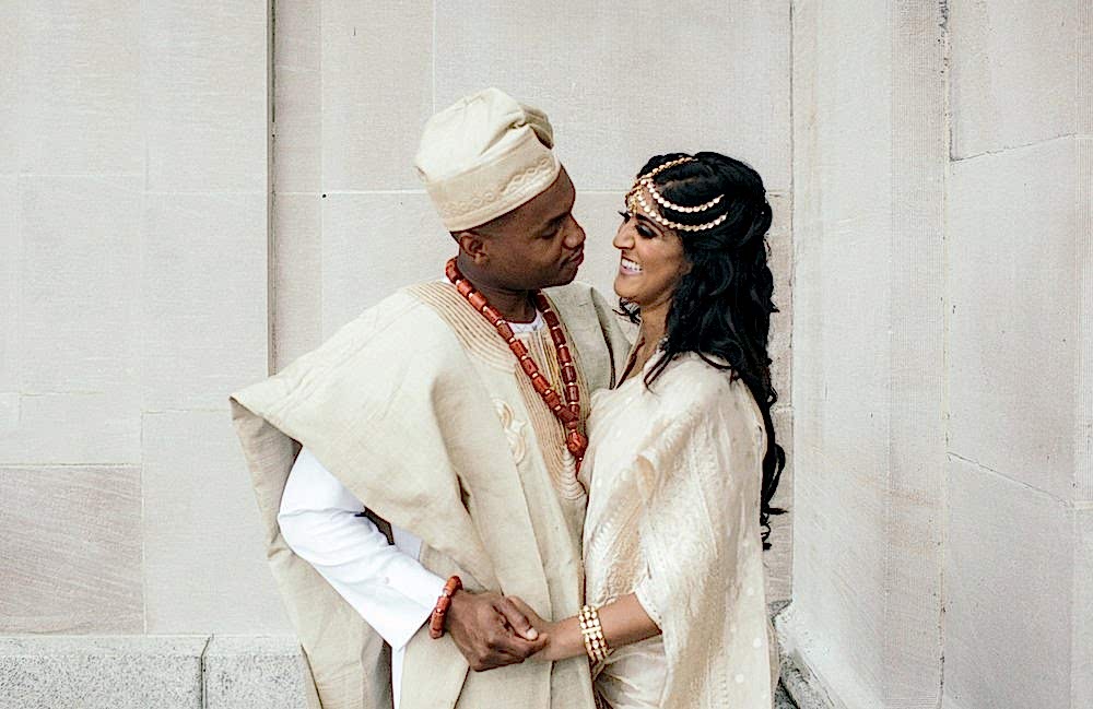A bride in a traditional Indian wedding outfit and a groom in traditional Nigerian wedding outfit hold hands and smile at each other in a photo by Betty Clicker