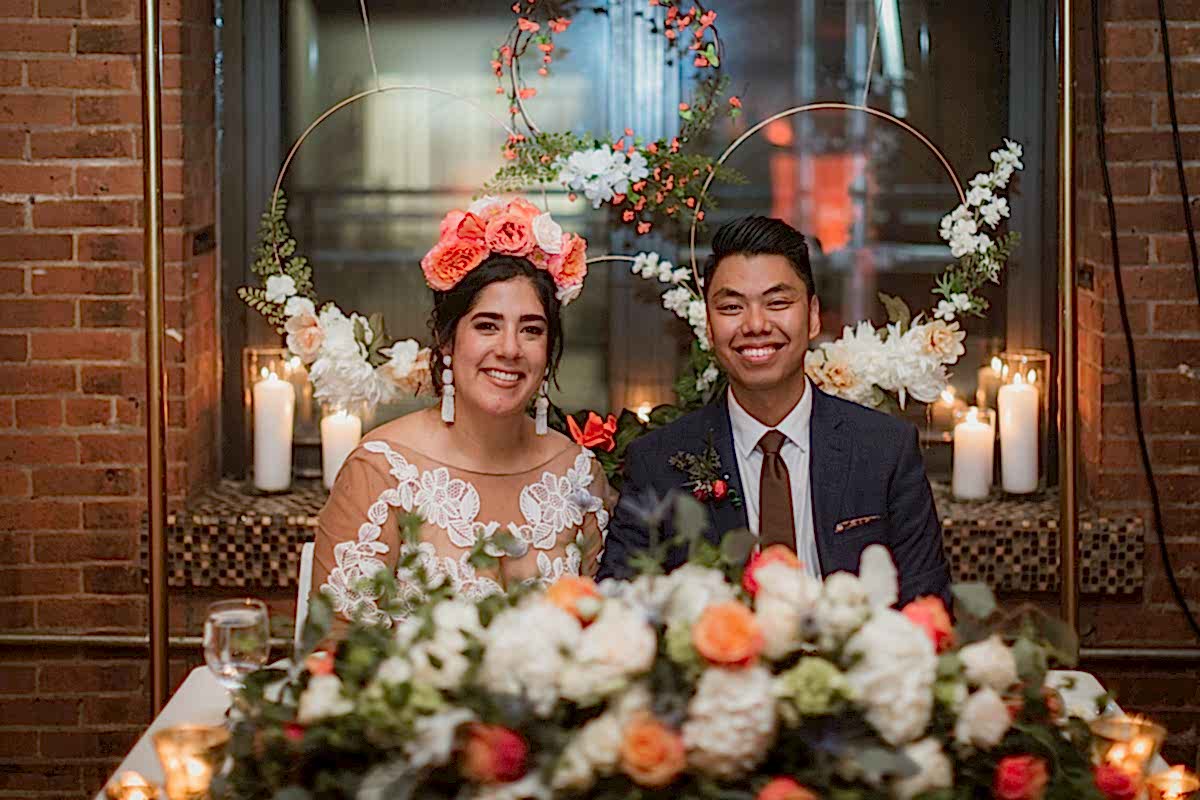 A bride wearing a crown of large salmon-colored flowers and a groom in a blue suit are seated at a sweetheart table behind a pile of matching flowers and look into the camera and smile in a photo by Betty Clicker