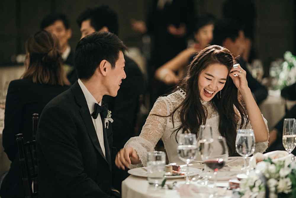 An Asian bride is sitting at a sweetheart table with her groom and she tucks her hair behind her ear as she laughs and reaches a hand out to groom in a photo by Betty Clicker