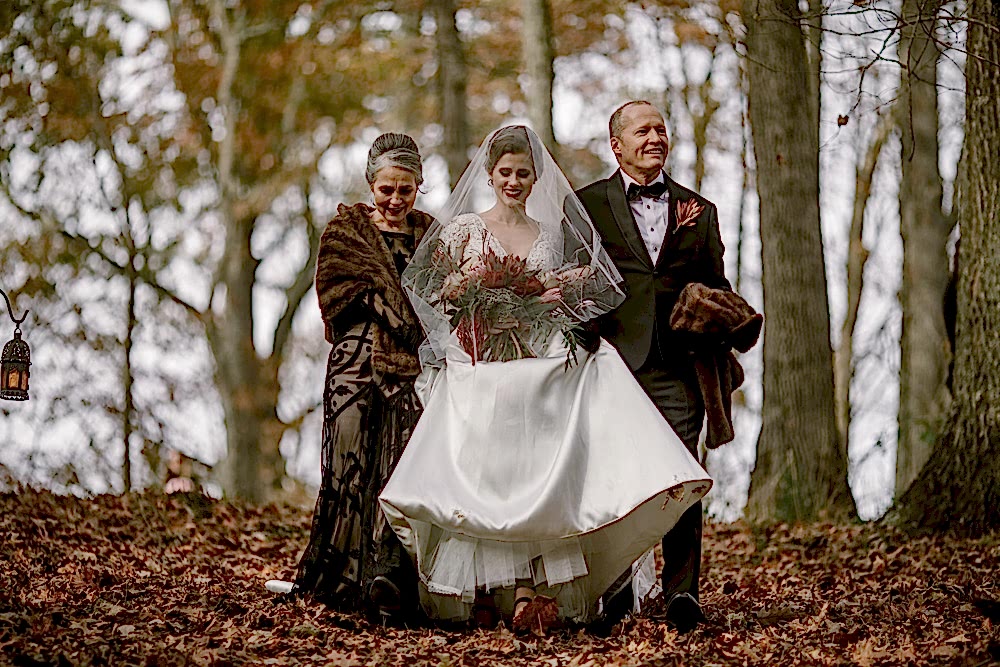 A bride wearing a veil is escorted down a hill covered in fall leaves with her mother in a brown gown and fur on her left, and her father in a suit on her right in a photo by Betty Clicker