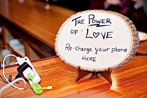 Wedding ideas for guest convenience—a charging station with a power strip and multiple adapters for phones next to a sign written on a slice of wood that reads, "The power of love... re-charge your phone here"