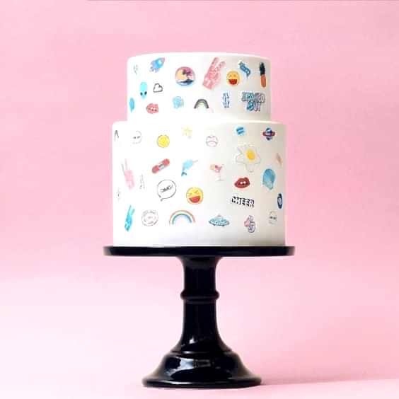 wedding ideas for cakes—two-tiered white cake with small emoji "stickers" adorning on a black pedestal stand