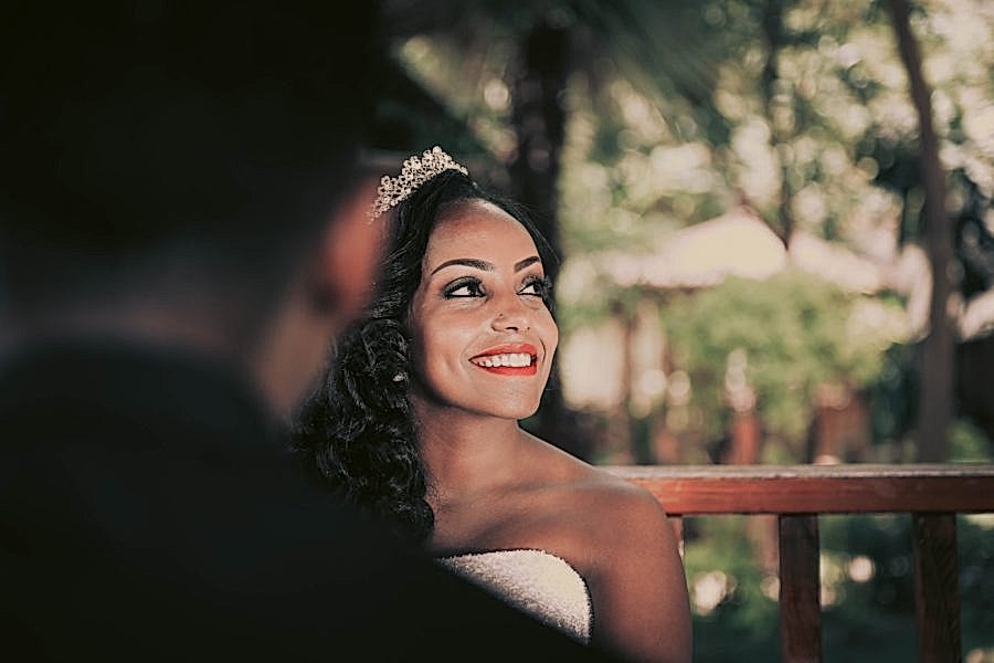 A woman with long wavy black hair wearing a tiara and wedding dress looks up, smiling
