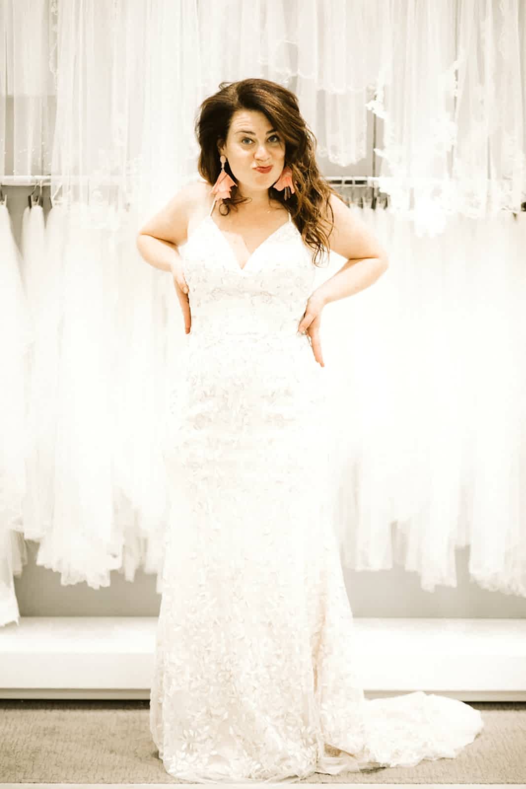 A woman with long brown hair stands with her hands on her hips and purses her lips while wearing a spaghetti-strap lace gown at David's Bridal