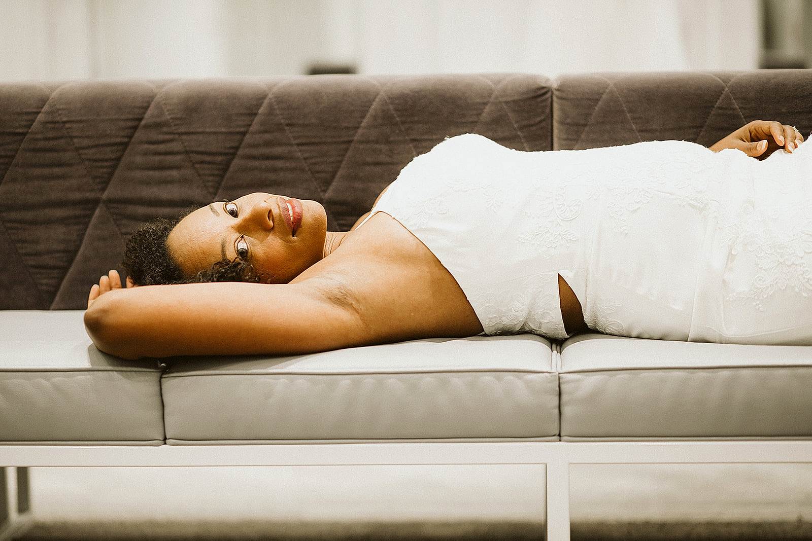 A woman with coiled dark hair lounges on a modern grey sofa and smiles slightly while looking at you as she wears a halter-neck white wedding dress in the fitting room at David's Bridal