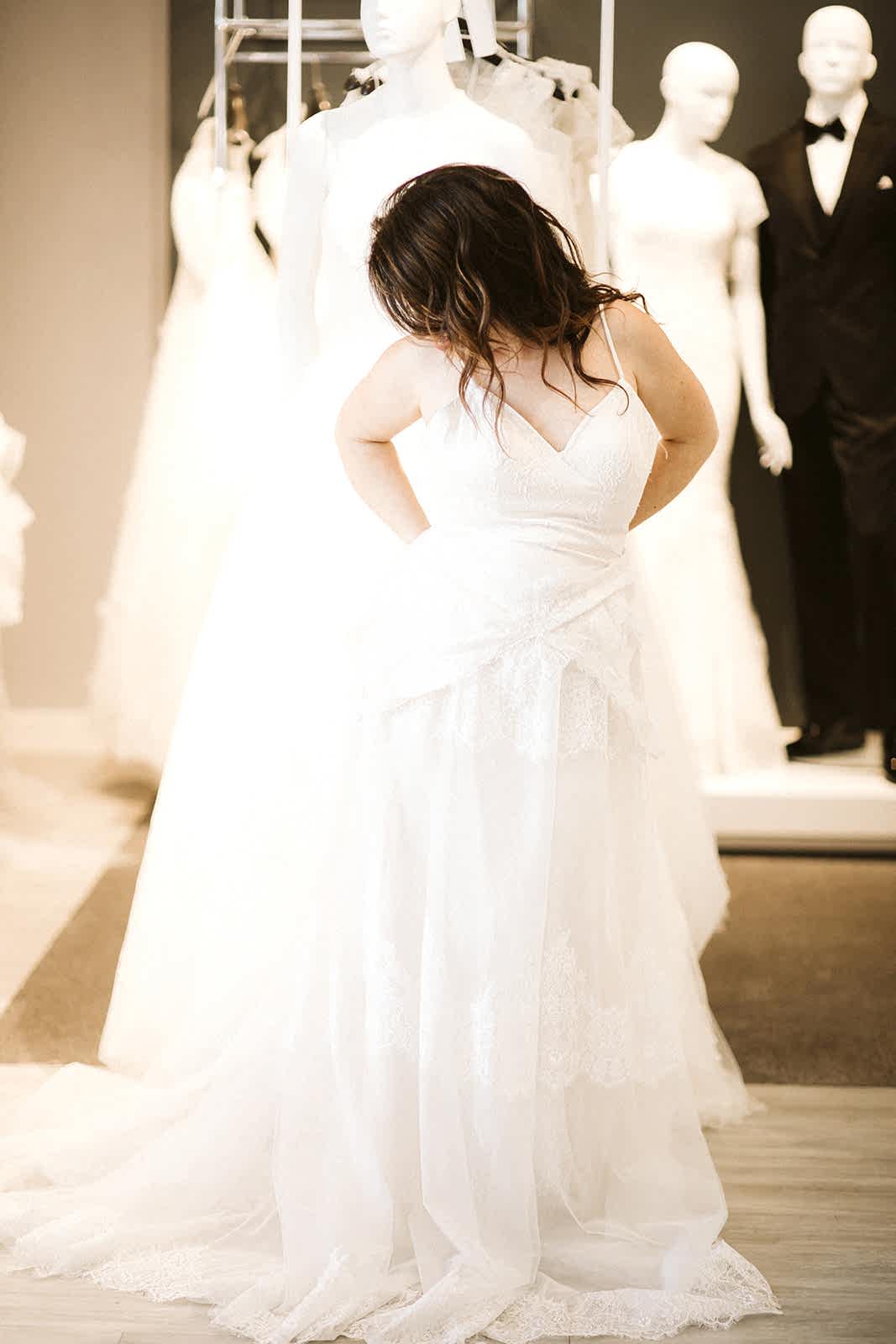 A woman with long brown hair begins to remove the outer dress (a sleeveless v-neck) to reveal a spaghetti-strap gown underneath at David's Bridal
