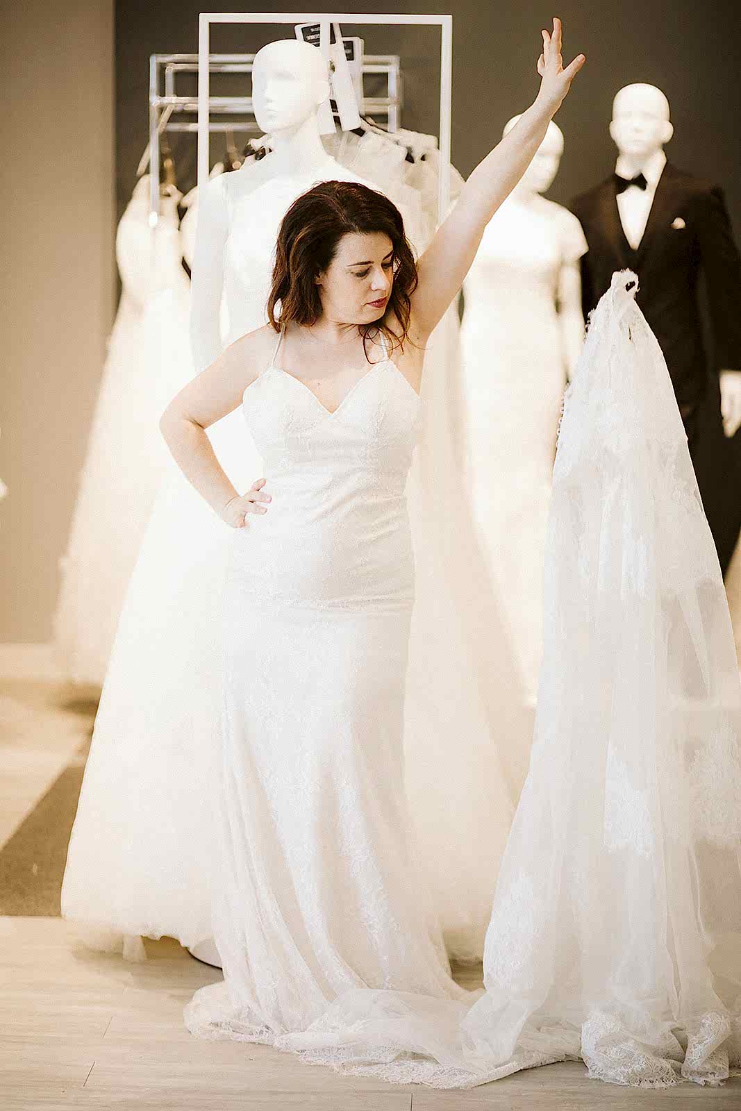 A woman with long brown hair stands with her hand on her hip as she drops the outer dress (a sleeveless v-neck) while wearing a spaghetti-strap gown at David's Bridal