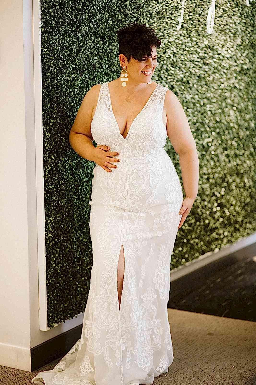 A curvy brunette woman wears a form-fitting sleeveless wedding gown with a plunge v-neckline and high center slit as she stands in front of a greenery wall at David's Bridal