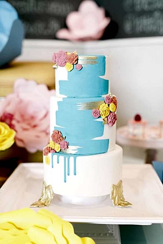 wedding ideas for cakes—a hand-painted cake with blue and silver streaks on three tier cake with gold leaf "feet" and pink, yellow, and orange edible rosettes 