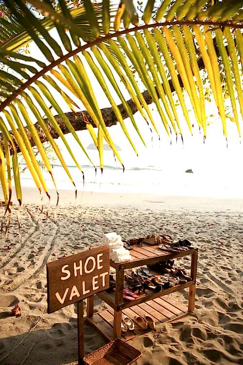 wedding ideas for a shoe valet on the beach—a sign reads "shoe valet" underneath a tropical tree on a sandy beach beside a three tiered shoe rack and small tower of rolled white hand towels