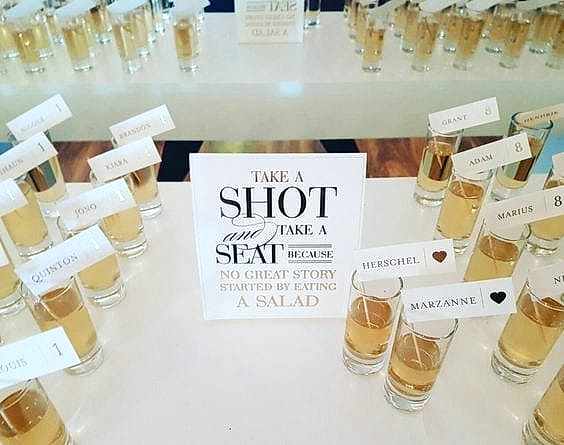 Wedding ideas for a seating chart—shots seating charts with a sign that reads "Take a shot and take a seat because no great story started by eating a salad" with rows of shot glasses with flags with names and table numbers