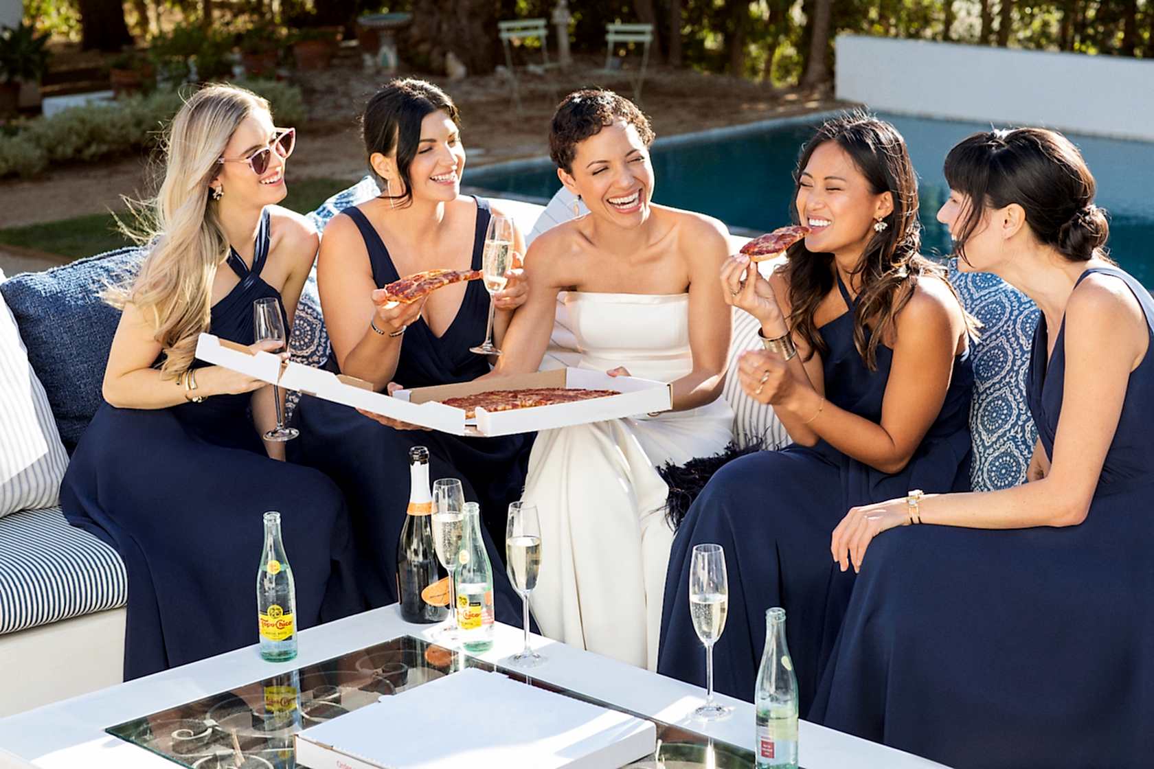 A bride and four bridesmaids in navy Brideside bridesmaid dresses eat pizza and drink champagne near an outdoor pool