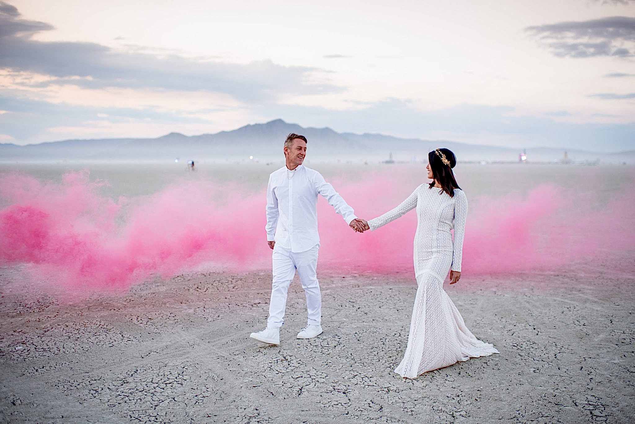 A man and a woman in white wedding clothes hold hands and look at each other as they walk in front of a pink smoke cloud in the desert.
