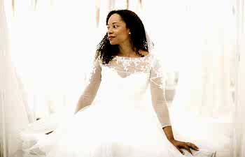 A woman sits in a bridal fitting room wearing a long sleeve wedding gown. She looks to one side, thoughtfully.