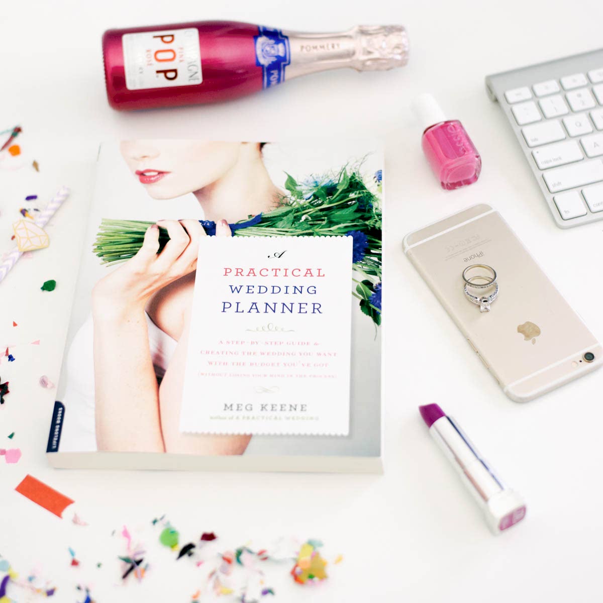 the a practical wedding planning guide on a white desk surrounded by champagne, makeup an iphone and confetti