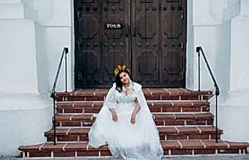 A woman in a radiant crown and a plus size wedding dress with a cape from Lace and Liberty x APW sits on terra cotta steps in front of brown wood doors