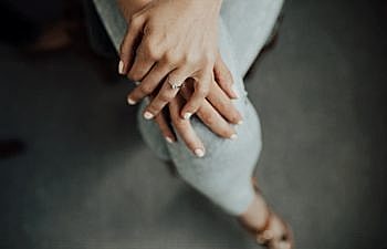 A woman in light grey leggings sits with her legs crossed, with her hands folded on her top knee, showing her diamond solitaire engagement ring.