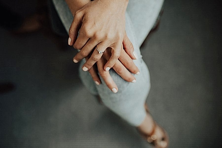 A woman in light grey leggings sits with her legs crossed, with her hands folded on her top knee, showing her diamond solitaire engagement ring.