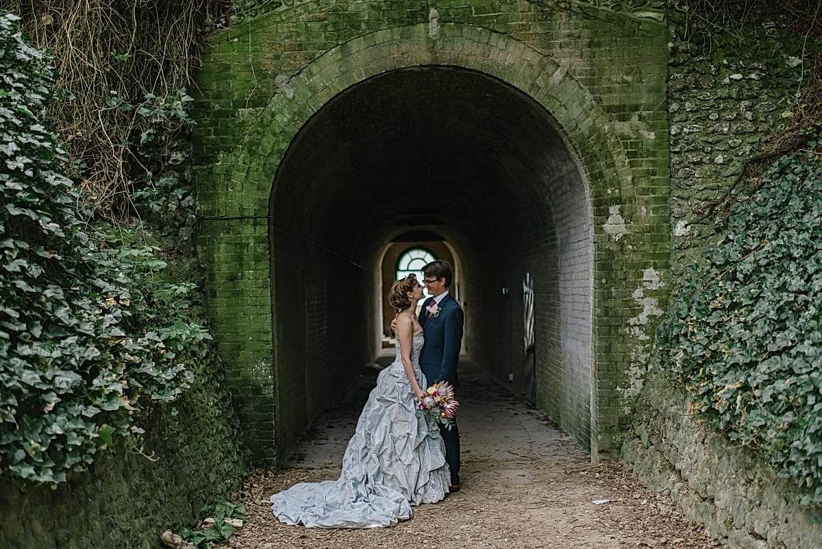 A couple in a wedding dress and tux stand at the moss- and ivy-covered opening of an old brick tunnel, smiling at each other at their destination wedding
