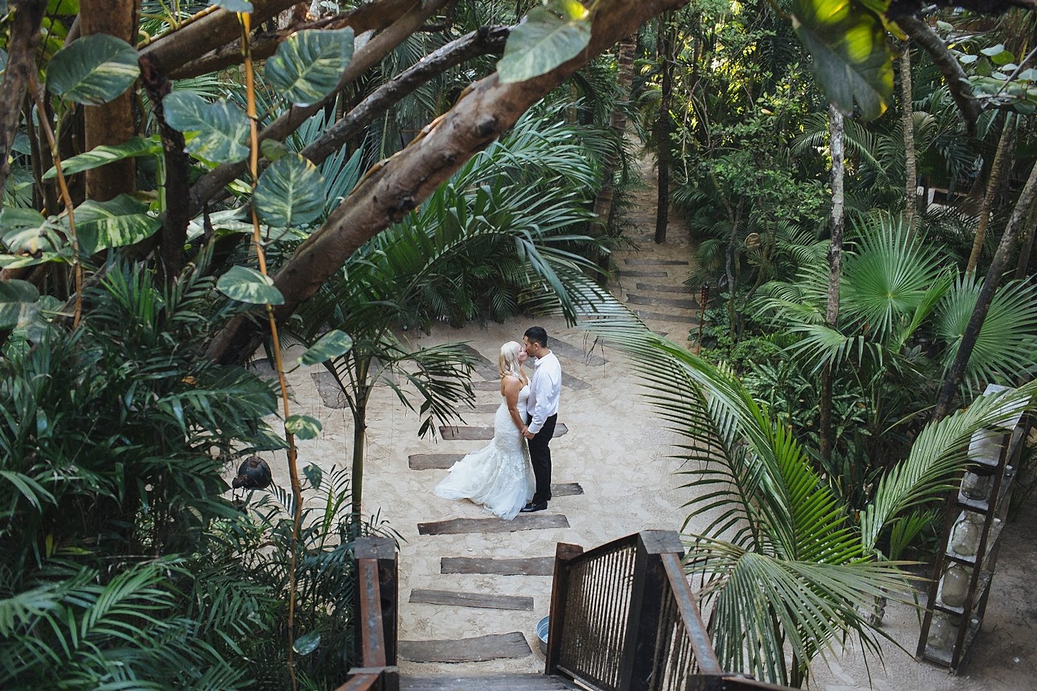 A couple in a wedding dress and shirt sleeves from a black tux stand amidst lush green tropical plants on a sandy path at their destination wedding
