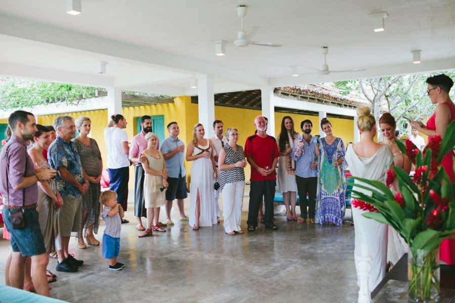 Two women in wedding dresses getting married at a tropical hotel, surrounded by family and friends standing in a semi-circle to witness the ceremony of their destination wedding.
