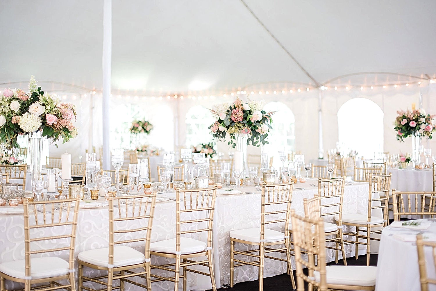 Gold Chiavari chairs at a white and blush tent wedding reception with raised flower arrangements on the tables.