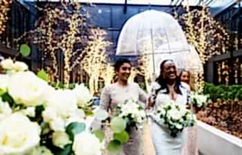 a bride and bridesmaid walk through twinkle lights while the bridesmaid carries a clear wedding umbrella for rain over the bride's head