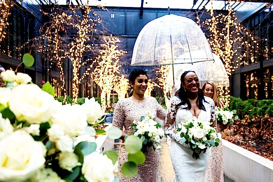 a bride and bridesmaid walk through twinkle lights while the bridesmaid carries a clear wedding umbrella for rain over the bride's head