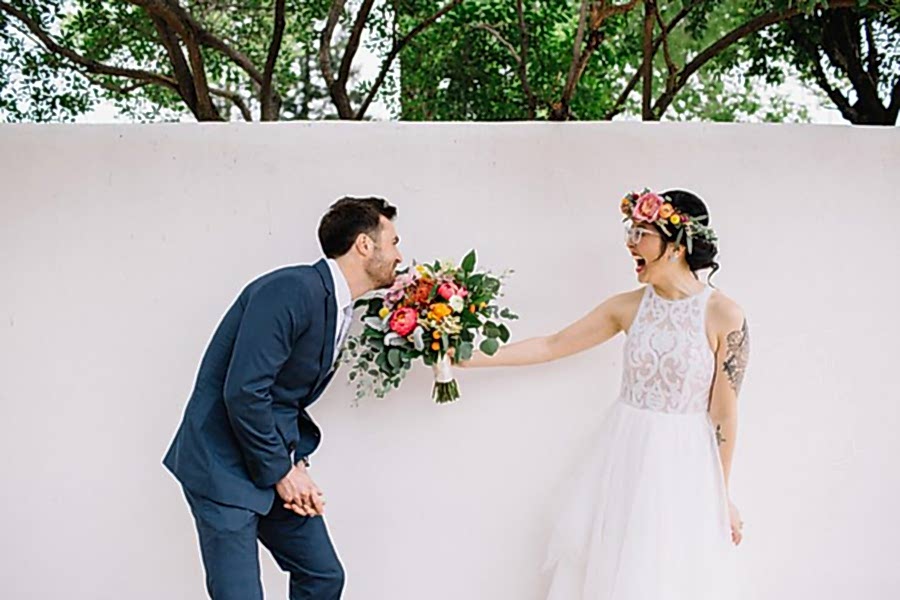 An Asian-American bride in a pink floral crown laughs as she holds out her bouquet for the groom to sniff