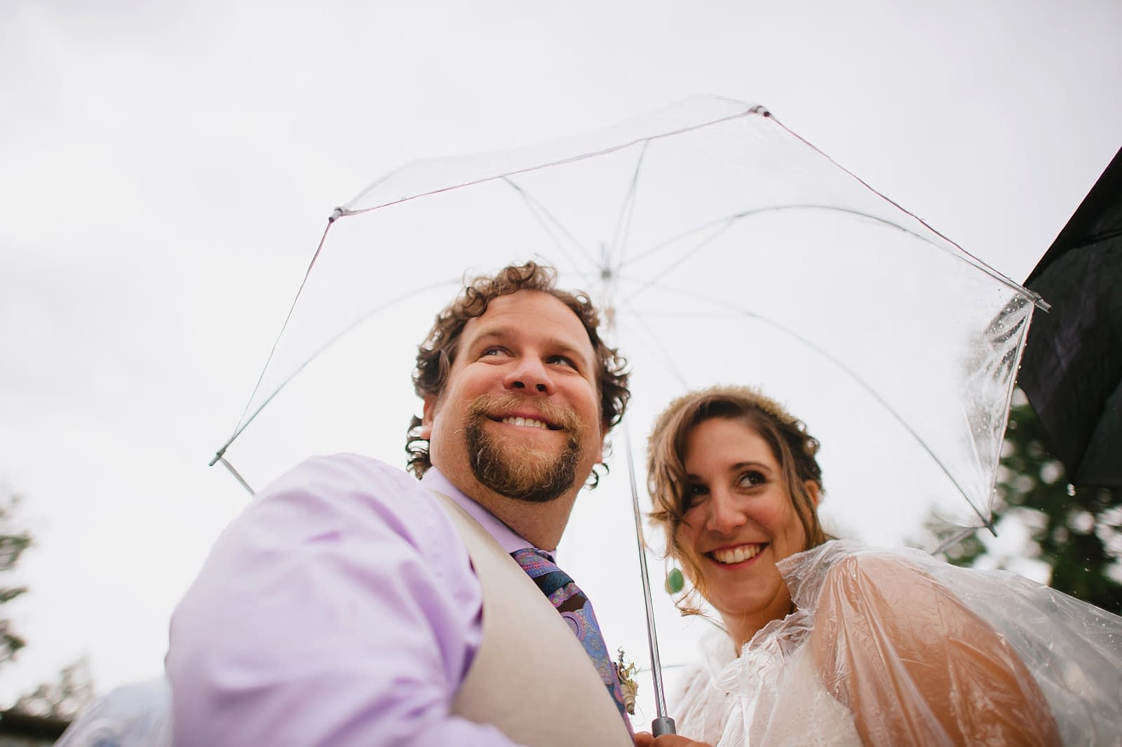 a couple smiling huddling under a wedding umbrella for rain while wearing a clear plastic rain ponchos