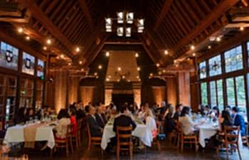 A large party gathers at several family-style tables in a large, high-windowed hall to celebrate an engagement party