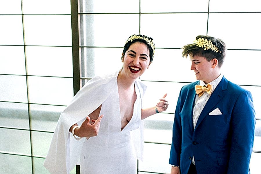 A queer couple in a wedding dress and a suit with bow tie. The person in a wedding dress laughs and points at themself, while the person in a suit smiles and looks on.