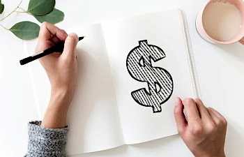 Hands holding a pen over a notebook with a large illustrated dollar sign drawn inside. The notebook sits next to a mug.